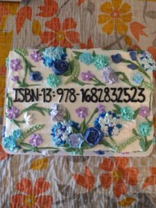 A rectangular cake with blue and purple flower decorations and the ISBN for "Lucky Bodies" written out in black frosting.